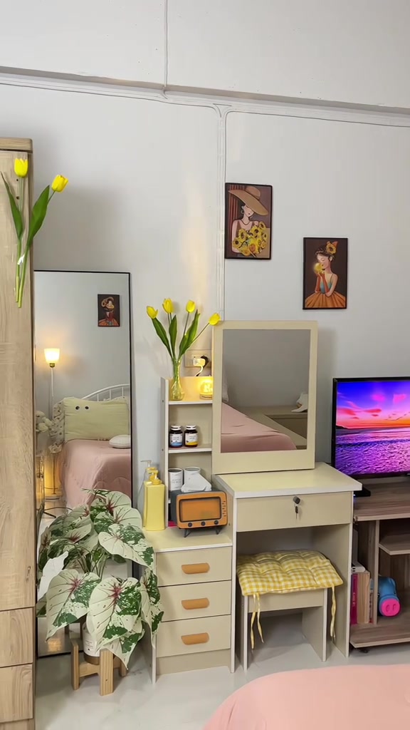 มุมห้องที่มีกระจกเงาบานเต็มตัวพิงข้างตู้ลิ้นชักสีครีม โต๊ะเครื่องแป้งกระจกเล็ก และทีวี