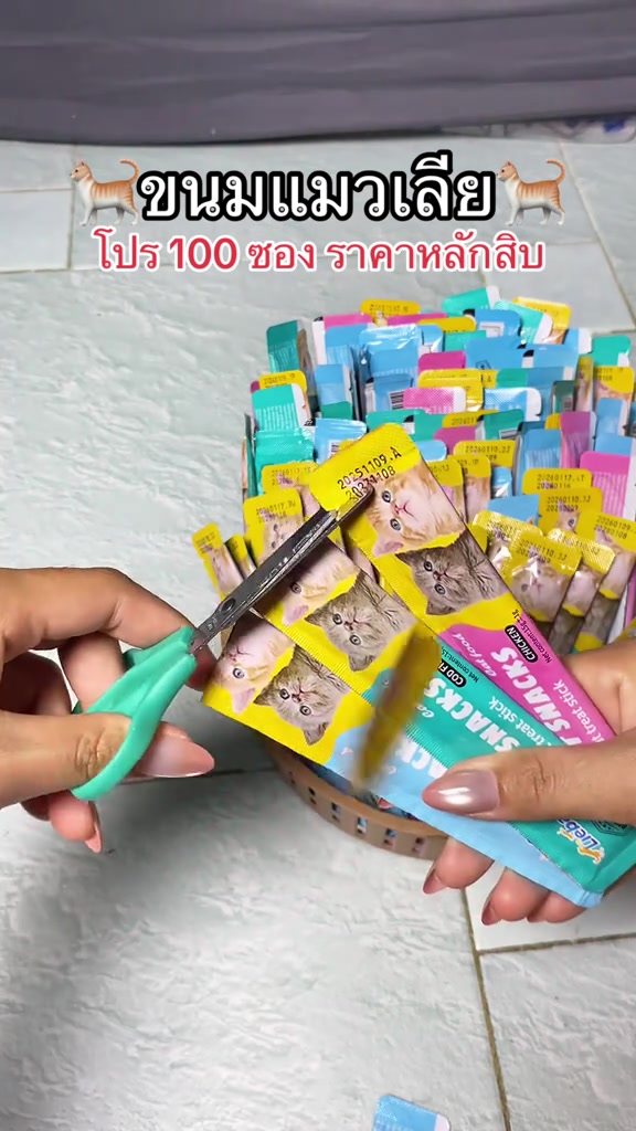 มือใช้กรรไกรตัดแถบซองขนมแมวเลียที่เชื่อมกัน โดยมีตะกร้าซองขนมหลากสีอยู่ด้านหลัง