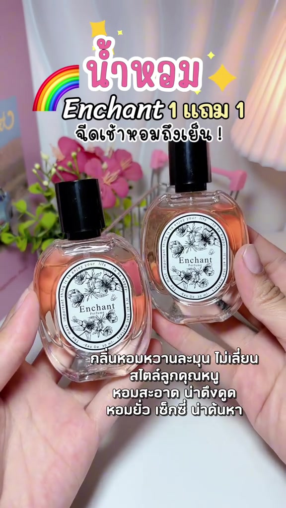 มือถือขวดน้ำหอมแก้ว Enchant Original ฉลากลายดอกไม้สองขวด พื้นหลังมีดอกสีชมพู