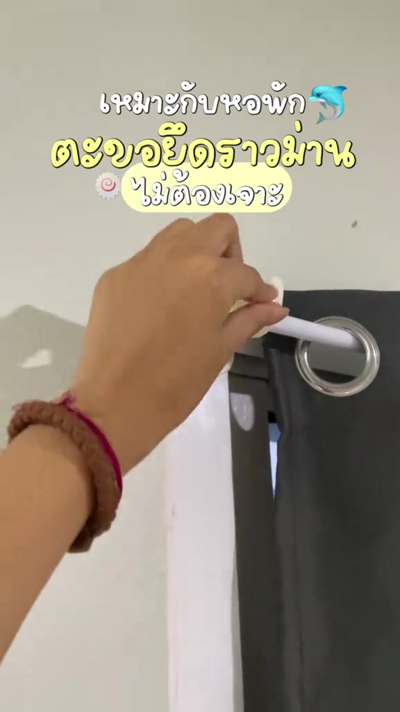 มือกำลังวางราวสีขาวลงบนตะขอยึดราวม่านแบบติดกาวเหนือม่านตาไก่สีเทา