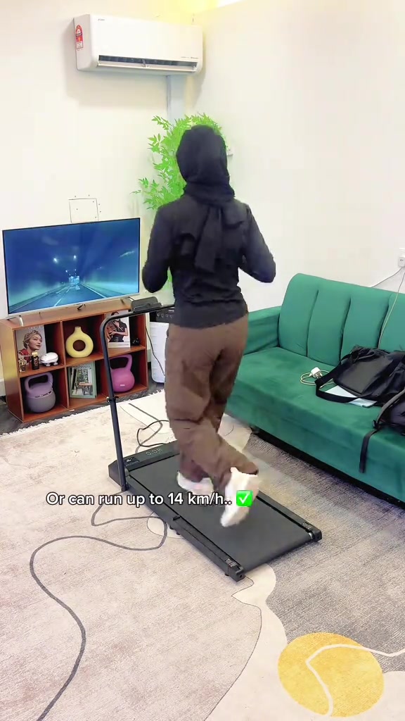 มุมมองด้านหลังผู้หญิงสวมเสื้อดำกำลัง jog บน walking pad ทรงเพรียวที่มีราวจับในห้องนั่งเล่นข้างโซฟาสีเขียว