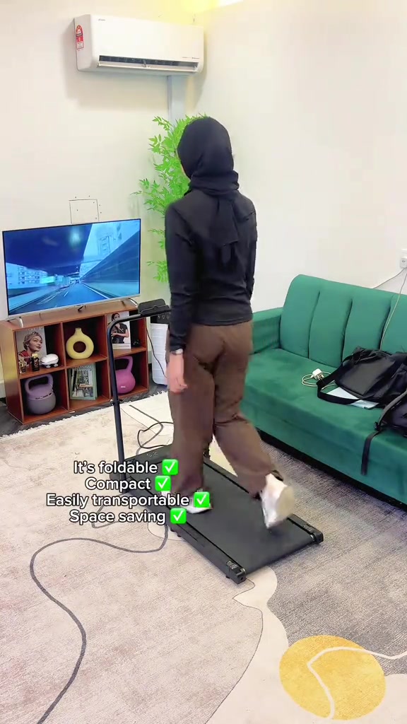 มุมมองด้านหลังผู้หญิงสวมเสื้อดำกางเกงน้ำตาลกำลังเดินบน walking pad ทรงเพรียวที่มีราวจับด้านหน้า วางบนพรมในห้องนั่งเล่นข้างโซฟาสีเขียว