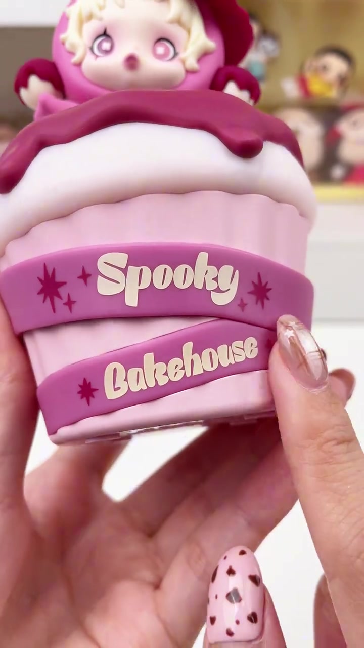 ภาพใกล้ของถ้วยเค้กชมพูแถบตัวอักษร Spooky Bakehouse และน้องหมวกแม่มดด้านบน