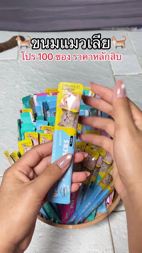 มือถือซองขนมแมวเลียสีฟ้าเหนือตะกร้าที่อัดแน่นด้วยซองขนมหลากสีนับร้อยซอง