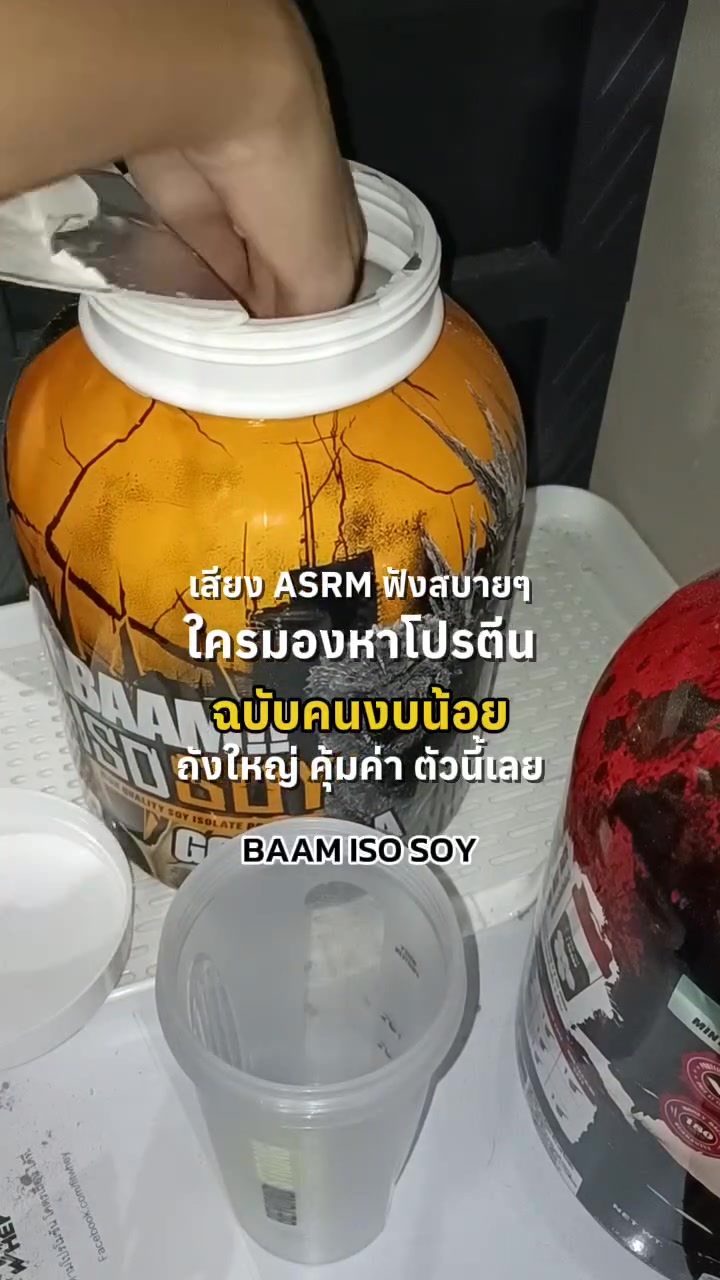 มือล้วงตักผงโปรตีนจากถัง BAAM ISO SOY สีส้มที่เปิดฝาแล้ว ด้านหน้ามีแก้วเชคใสวางอยู่