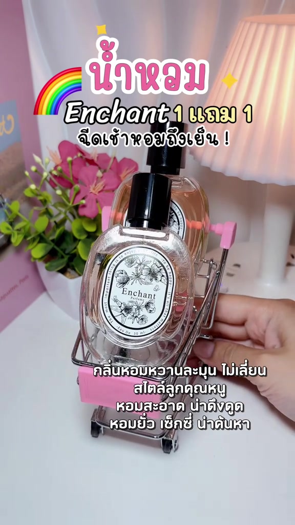 ขวดน้ำหอม Enchant สองขวดวางในรถเข็นมินิสีชมพูบนโต๊ะสีขาวข้างโคมไฟ