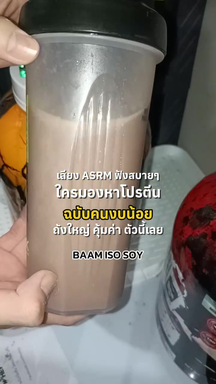 มือถือแก้วเชคโปรตีนสีน้ำตาลอ่อนที่ชงเสร็จแล้วข้างถังโปรตีน