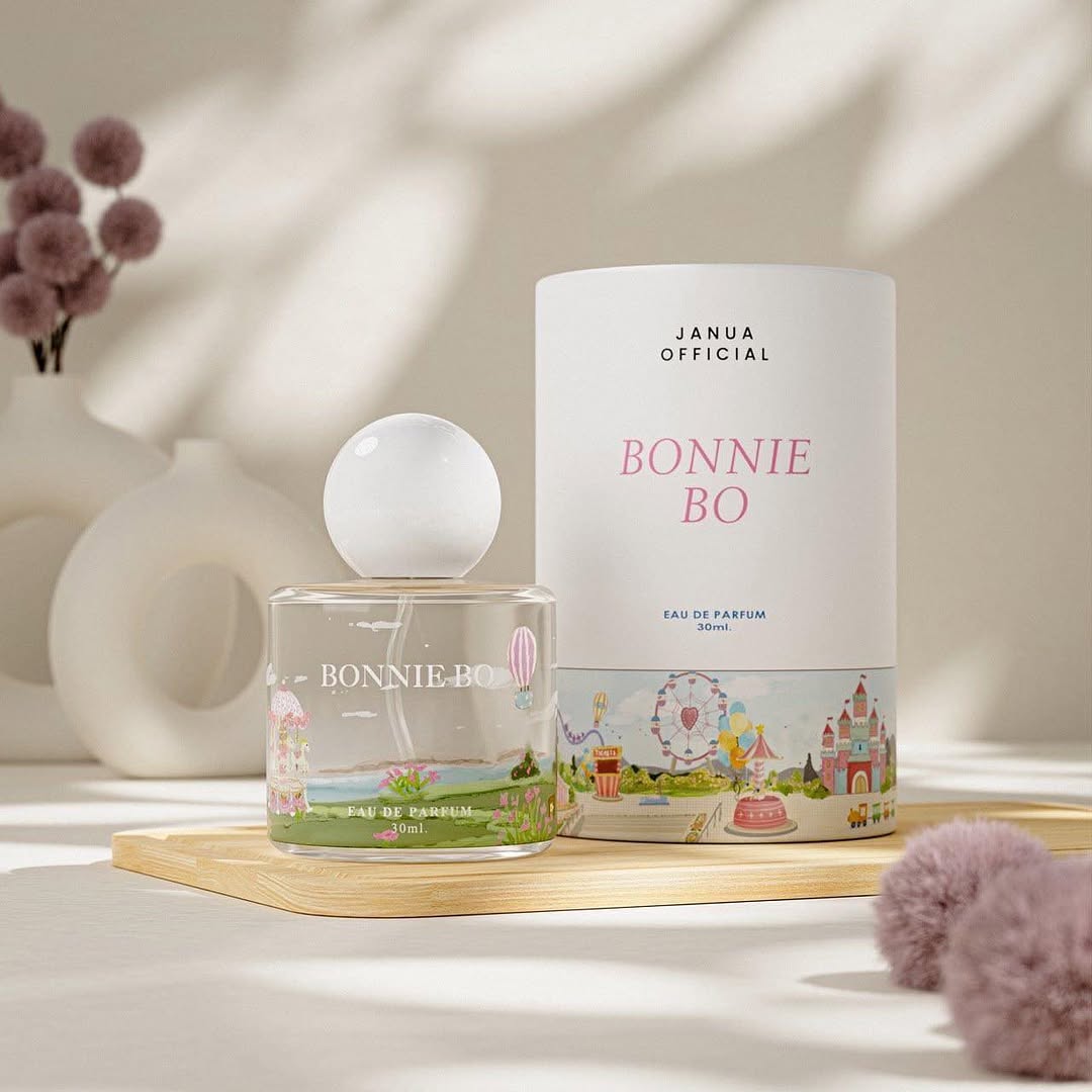 ขวดน้ำหอม Janua Bonnie Bo ฝาทรงกลมสีขาววางคู่กล่องทรงกระบอกพิมพ์ลายสวนสนุกบนถาดไม้