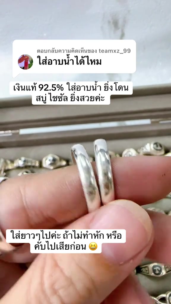 นิ้วถือแหวนเงินแท้ทรงเกลี้ยงสองวงคู่กัน พื้นหลังเป็นถาดใส่แหวนเงินเรียงหลายวง
