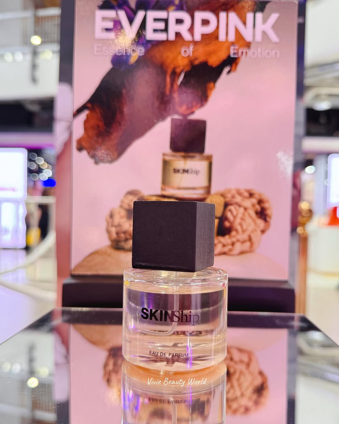 น้ำหอม EVERPINK Skinship ขวดแก้วใสฝาสี่เหลี่ยมสีดำ ตั้งบนโต๊ะกระจกเงา ด้านหลังเป็นโพสเตอร์สีชมพูของคอลเลกชัน