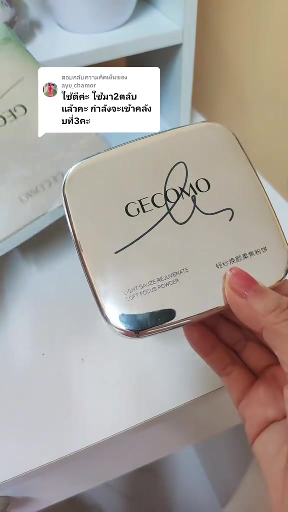 มือถือตลับแป้งจีโคโมฝาเงินมันวาวโลโก้สีดำ บนพื้นหลังชั้นวางเครื่องสำอางสีขาว