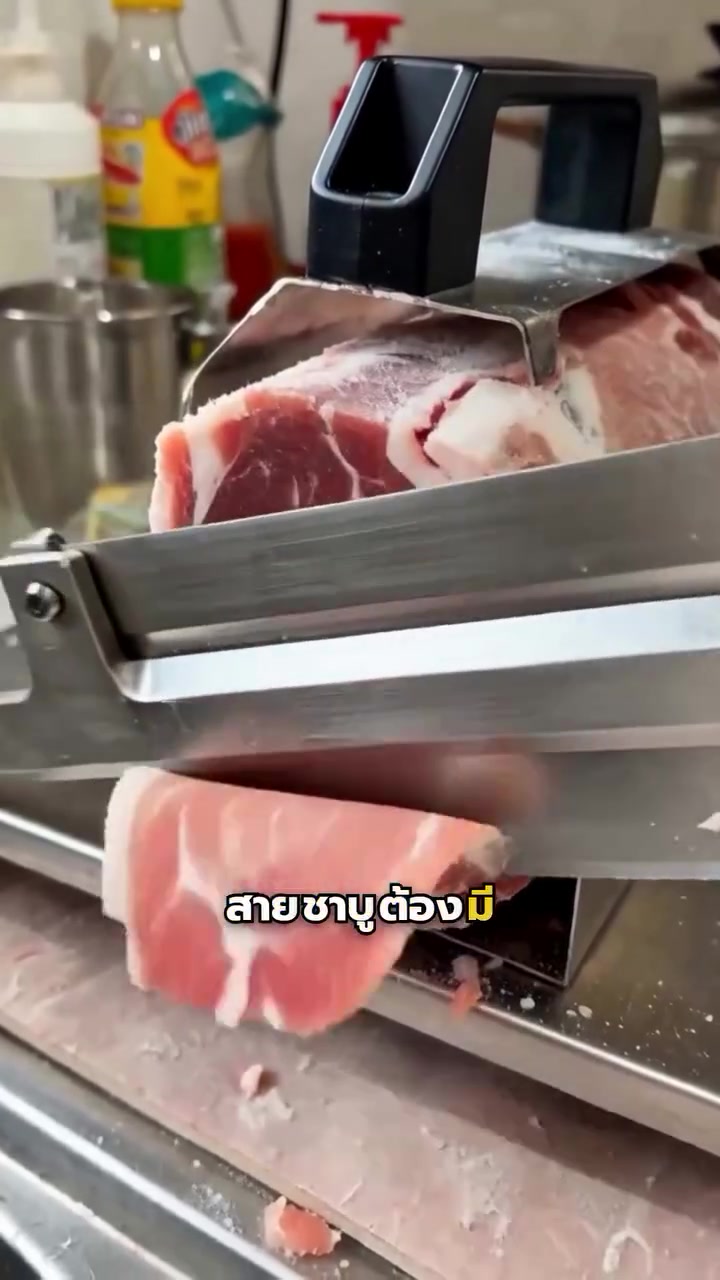 ก้อนหมูวางอยู่ในแท่นกดของที่สไลด์เนื้อ ใบมีดดันแผ่นหมูบางสีชมพูออกมาด้านข้าง