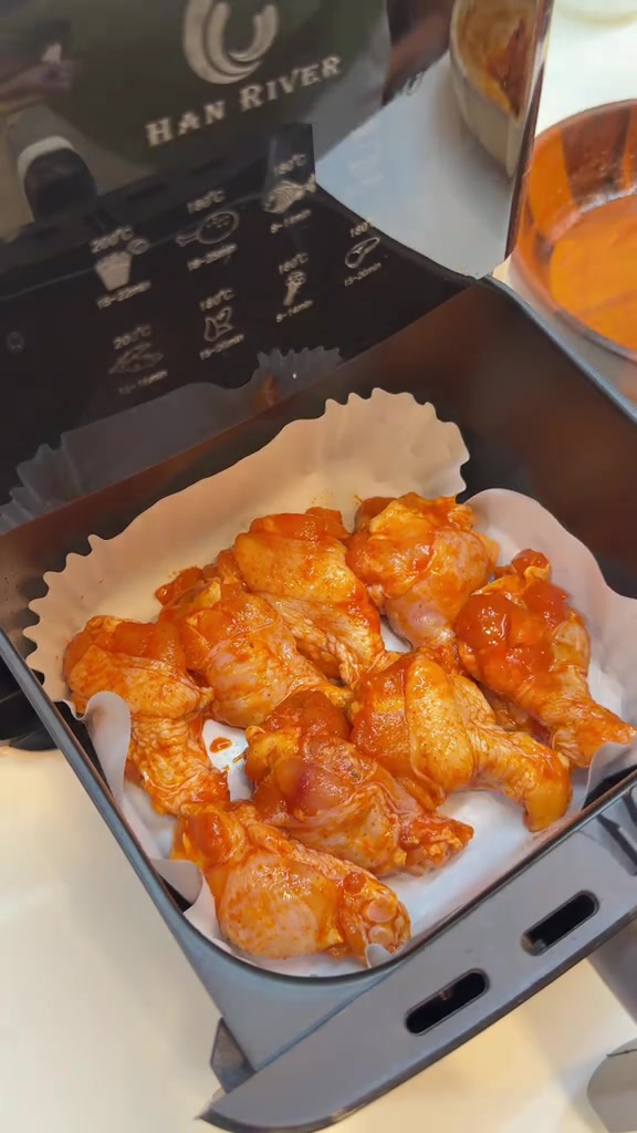 ปีกไก่หมักซอสเรียงอยู่บนกระดาษรองทรงกลมในตะกร้าหม้อทอดไร้น้ำมัน