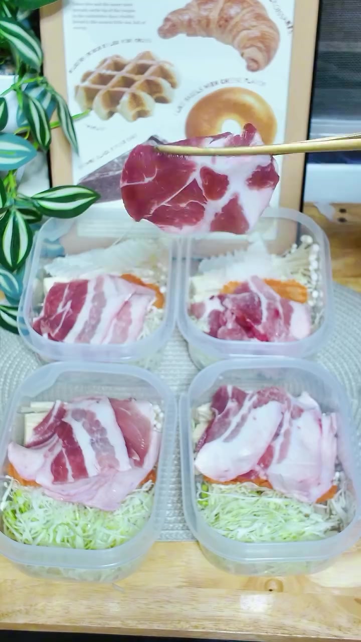 ตะเกียบคีบหมูสไลด์แผ่นบางลอยเหนือกล่องถนอมอาหารสี่กล่องที่จัดหมูและผักซอยไว้เป็นชุดมื้อๆ