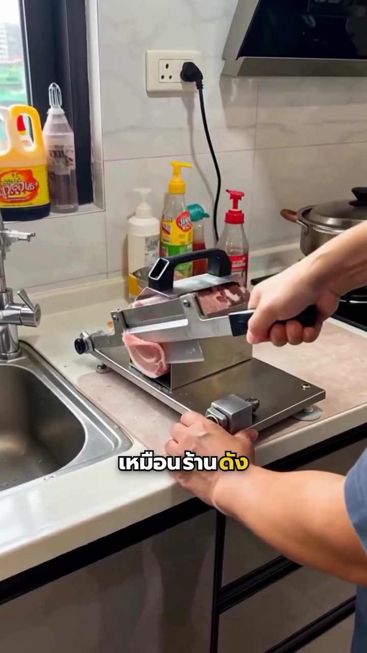 ที่สไลด์เนื้อแบบมือโยกตัวสแตนเลสวางบนเคาน์เตอร์ครัวในบ้าน มือหนึ่งจับด้ามโยกใบมีด อีกมือประคองฐานเครื่องไว้