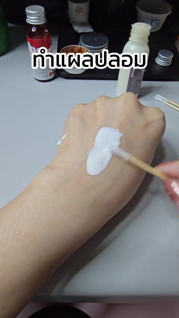 มือใช้ไม้เล็กทาครีมสีขาวเป็นปื้นบนหลังมือ โดยบนโต๊ะมีขวดเลือดปลอมและขวดน้ำยางสีขาววางอยู่