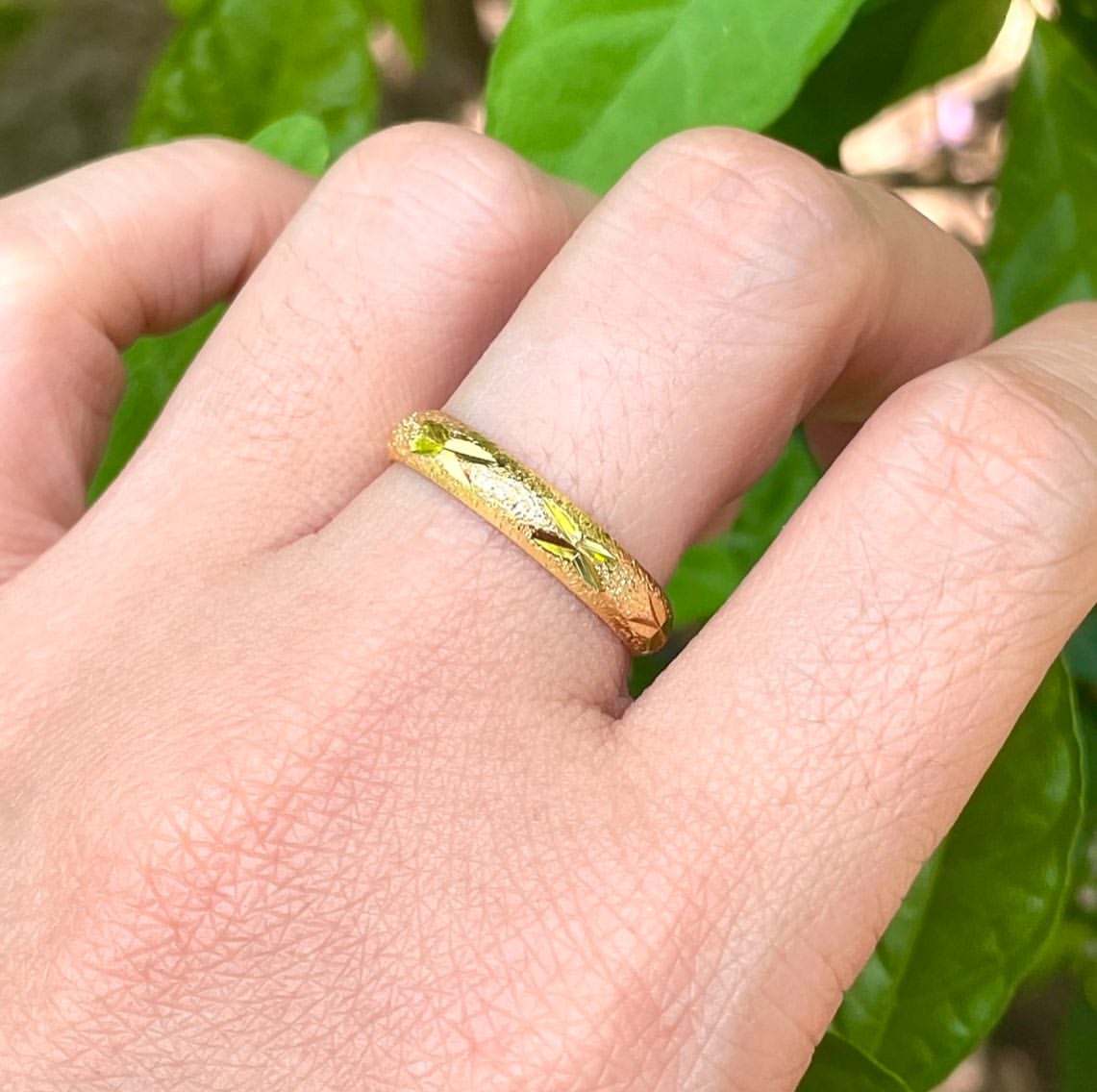 แหวนทองไมครอนวงเรียวลายดาวสวมบนนิ้ว มือวางบนพื้นหลังใบไม้สีเขียว