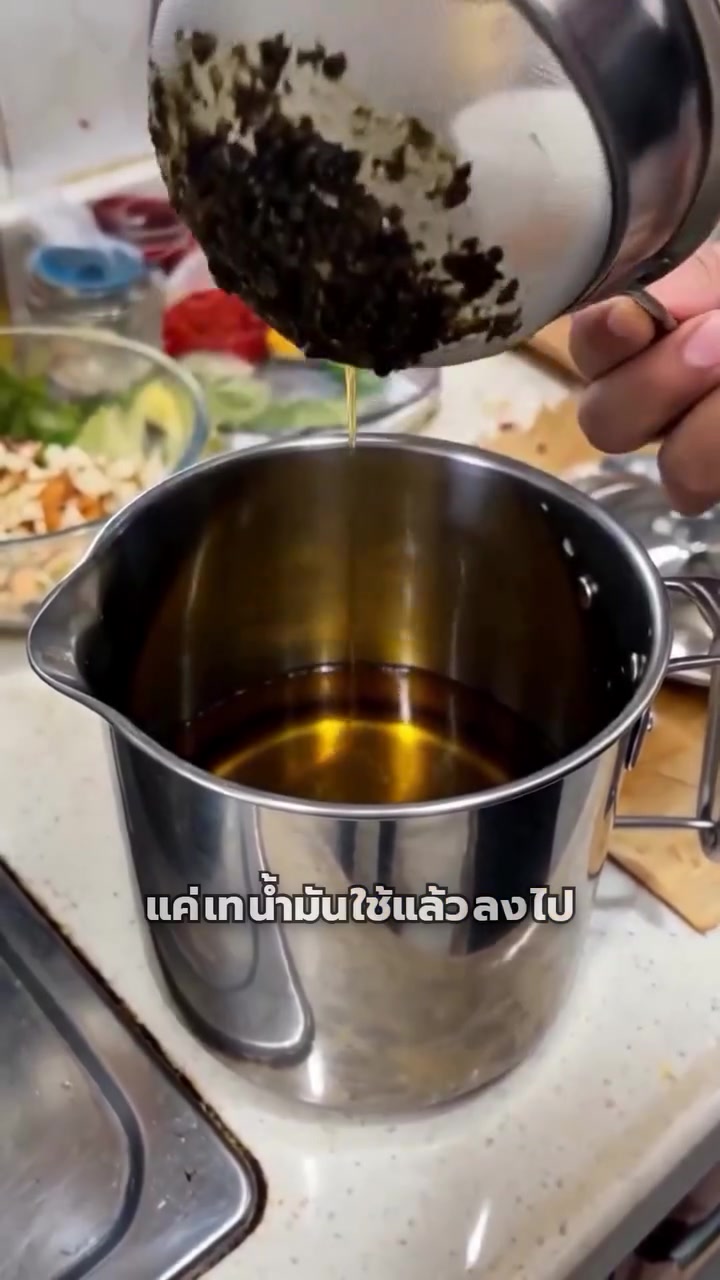 ยกตะแกรงกรองที่มีเศษทอดไหม้ติดอยู่ออกจากปากหม้อ น้ำมันใสไหลเป็นสายลงสู่หม้อสแตนเลสด้านล่าง