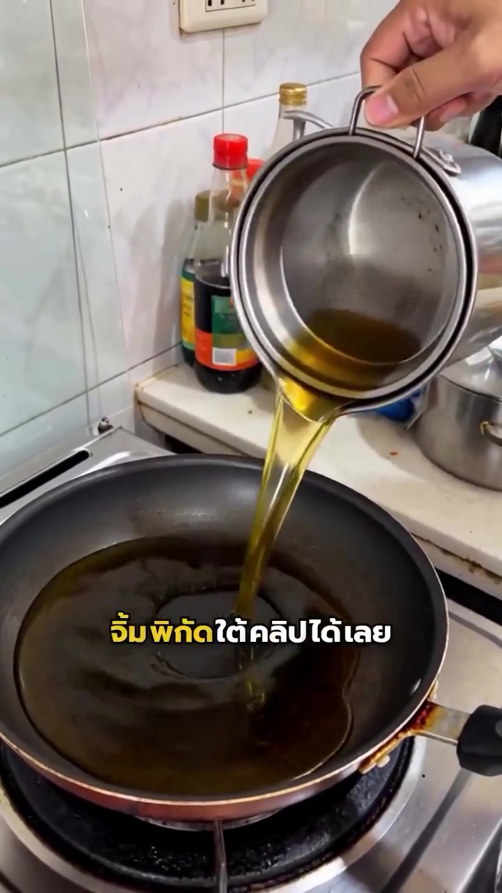 เทน้ำมันที่กรองแล้วจากหม้อสแตนเลสผ่านตะแกรงตาถี่ลงกระทะบนเตาเพื่อใช้ทอดซ้ำ