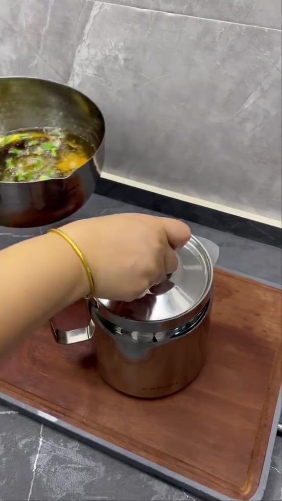 มือกำลังปิดฝาสแตนเลสของหม้อกรองน้ำมันที่วางบนเขียงไม้ โดยมีกระทะใส่น้ำมันทอดใช้แล้วอยู่ด้านหลัง