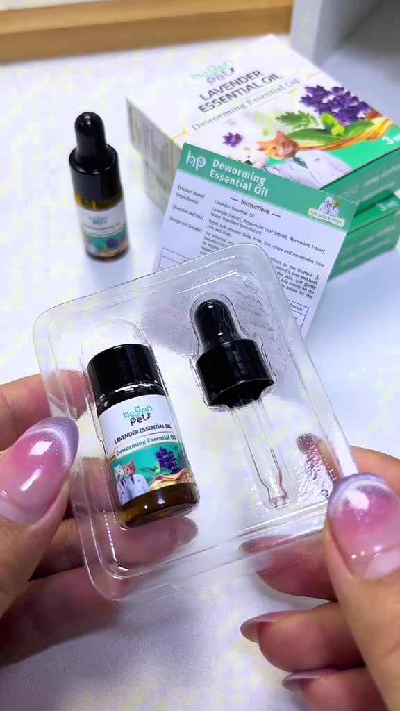 มือถือถาดพลาสติกใสบรรจุขวดน้ำมันลาเวนเดอร์สีชาพร้อมหลอดหยด วางคู่กล่องกระดาษลายดอกลาเวนเดอร์
