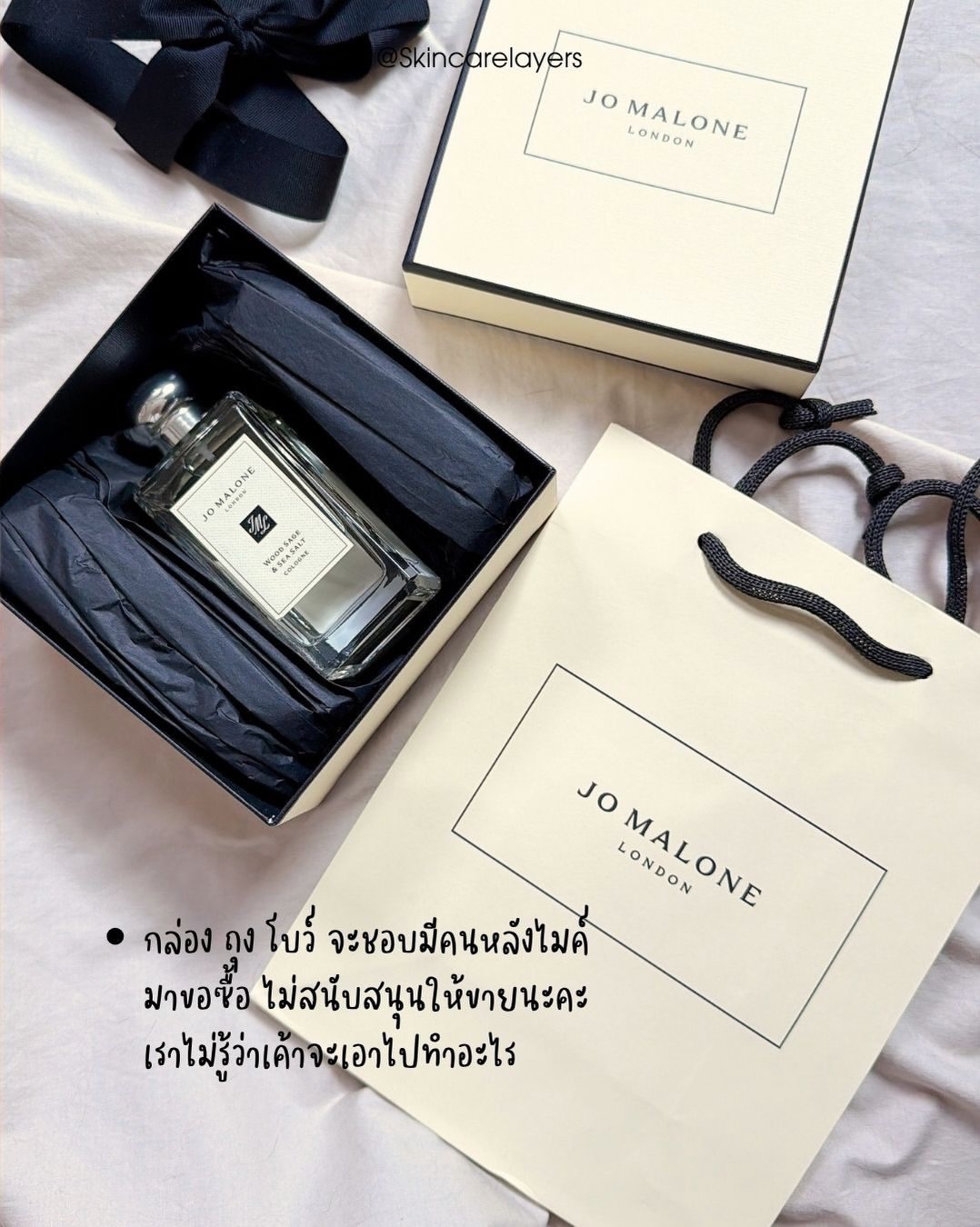 ขวด Jo Malone Wood Sage & Sea Salt ในกล่องสีดำวางคู่ถุงกระดาษสีครีมของแบรนด์บนผ้าสีขาว