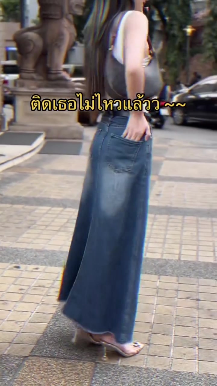 ด้านข้างของสาวสวมเสื้อสีขาวกับกระโปรงยีนส์ยาวสีเข้ม สะพายกระเป๋าใบเล็กบนทางเท้า