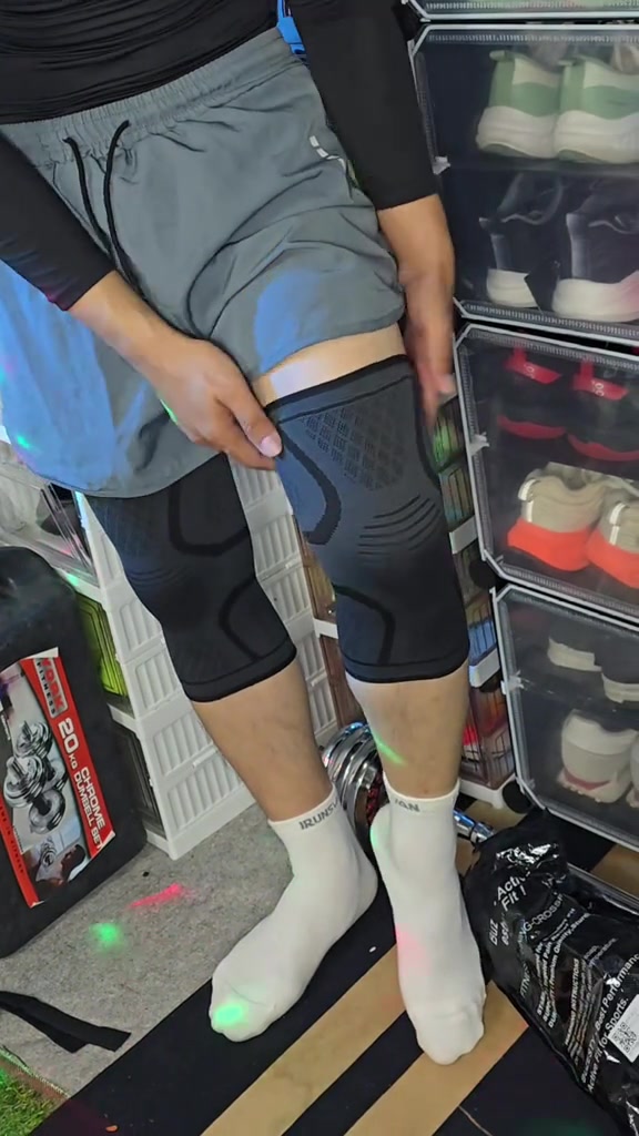 คนยืนสวมปลอกเข่าผ้าถักสีเทาดำทั้งสองข้าง มือจับขอบปลอกด้านบนเช็กความกระชับ ฉากหลังเป็นชั้นวางรองเท้าในบ้าน