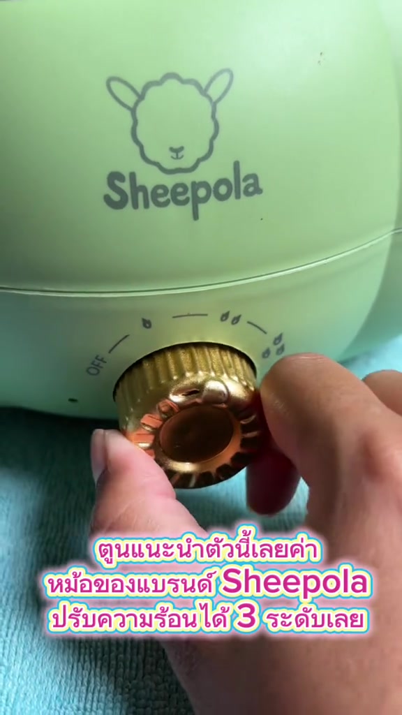 มือกำลังหมุนปุ่มปรับความร้อนสีทองบนตัวหม้อสีเขียว เห็นเครื่องหมายปิดและระดับไฟสามระดับพร้อมโลโก้ Sheepola เหนือปุ่ม