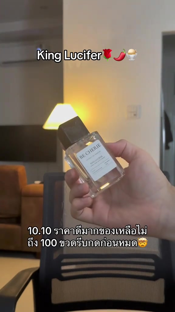 มือถือขวดน้ำหอม BE CHERIE ฝาดำเอียงหน้าฉากขาวให้กล้อง ฉากหลังเป็นโคมไฟแสงอุ่นในห้องนั่งเล่น