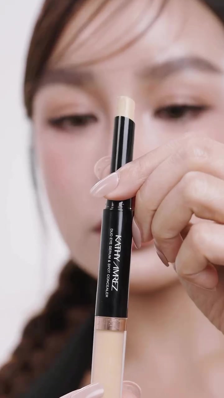 มือถือแท่งคอนซีลเลอร์สองหัวแคธี อัมเรซลำกล้องดำโดยเปิดฝั่งสติ๊กสีเบจด้านบนและเห็นฝั่งเซรั่มด้านล่าง