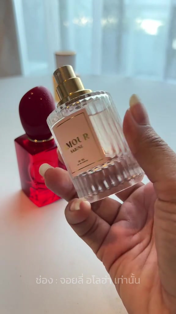 มือถือขวด AMOUR Sabine เปิดฝาเห็นหัวฉีดทอง วางเทียบข้างขวดน้ำหอมสีแดงของ Giorgio Armani Si Passione บนโต๊ะขาว