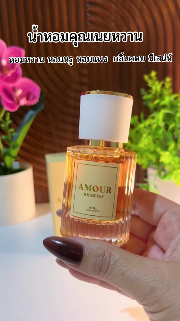 มือถือขวดน้ำหอม AMOUR Richesse ขวดแก้วใสมีลายร่อง น้ำหอมสีอำพัน ฝาขาวขอบทอง ฉลากครีมอ่านว่า AMOUR RICHESSE 30 ML ฉากหลังเป็นกล้วยไม้สีชมพูและผนังไม้