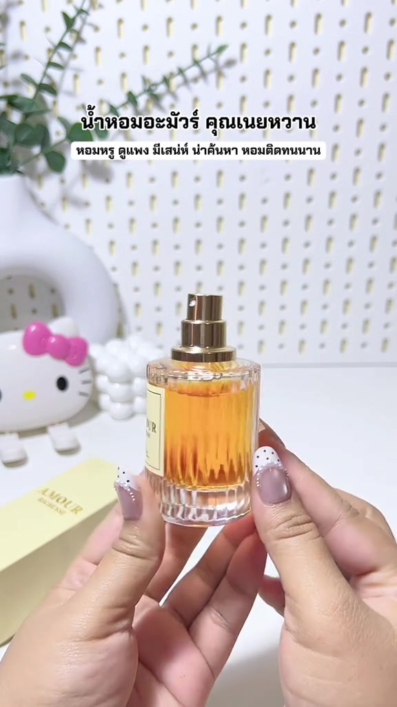 มือสองข้างถือขวด AMOUR Richesse แบบเปิดฝา เห็นหัวฉีดสเปรย์สีทองและน้ำหอมสีอำพันภายในขวดแก้วลายร่อง ฉากหลังเป็นโต๊ะขาวและตุ๊กตา Hello Kitty