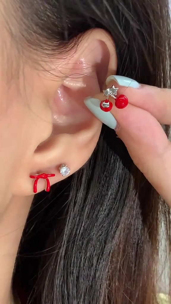 มือเล็บสีมินต์ถือจิวแป้นแบนหัวเชอร์รีแดงจ่อที่ใบหู บนติ่งหูมีจิวทรงริบบิ้นแดงและหัวเพชรเล็กใส่อยู่