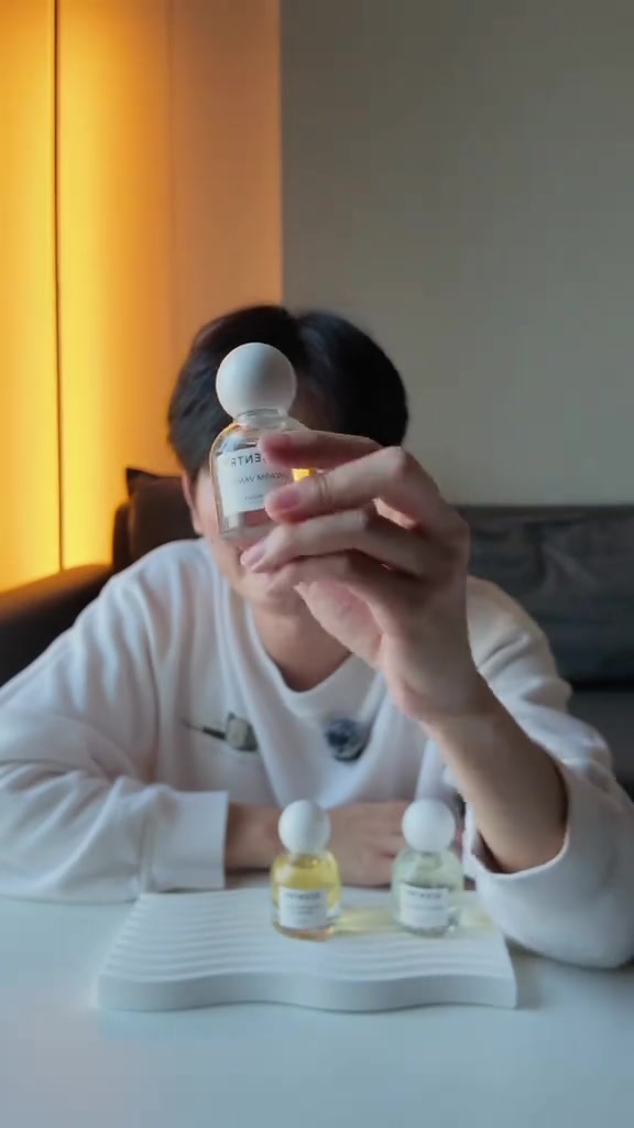 มือชูขวดน้ำหอม Scentry ฝากลมสีขาวเหนือถาดเซรามิกสีขาวลอนคลื่นที่มีอีกสองขวดวางอยู่