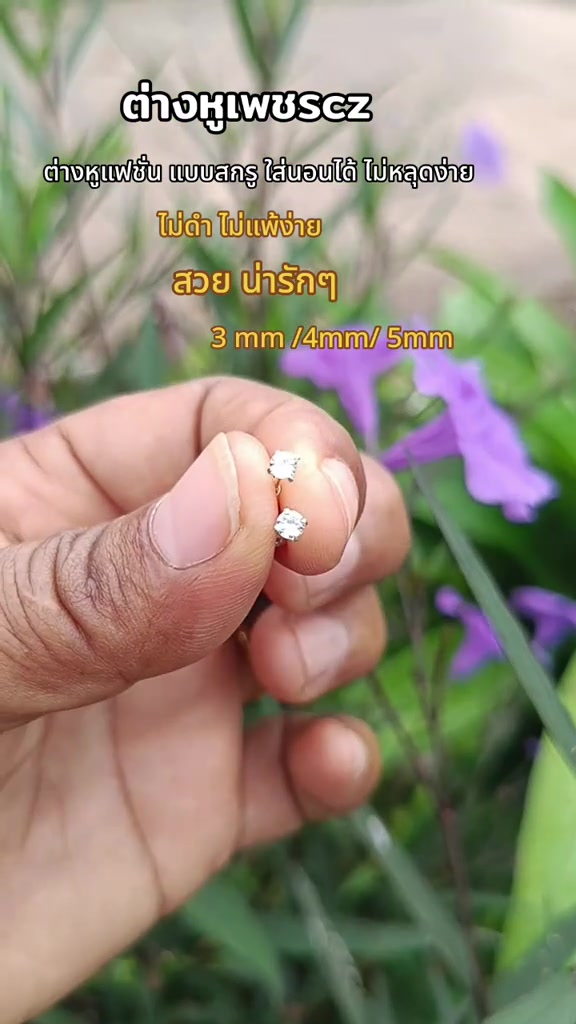 นิ้วมือหนีบต่างหูเพชรซีแซดทรงกลมสองเม็ดระยะใกล้ เห็นเหลี่ยมหินสะท้อนแสงชัดบนพื้นหลังดอกไม้สีม่วง