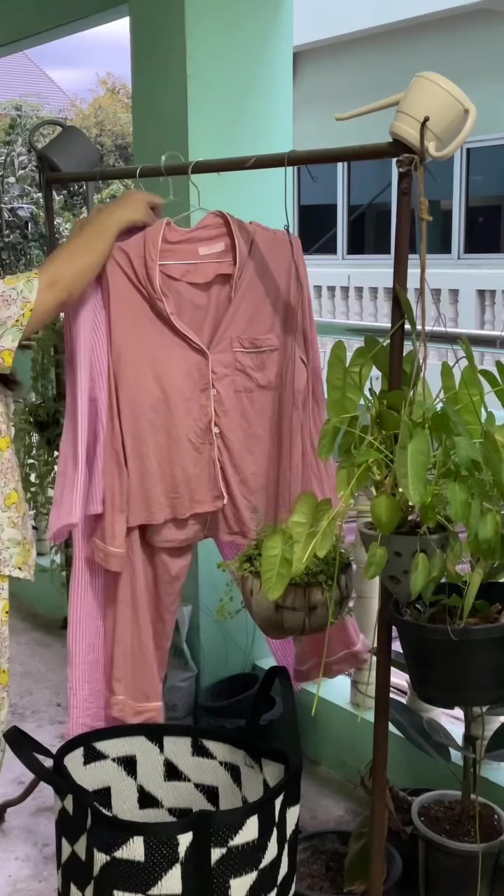 ชุดนอนสีชมพูตากบนราวเหล็กที่ระเบียงห้อง มือกำลังแขวนไม้แขวนเสื้อขึ้นราว ข้างราวมีกระถางต้นไม้แขวนเรียงกัน