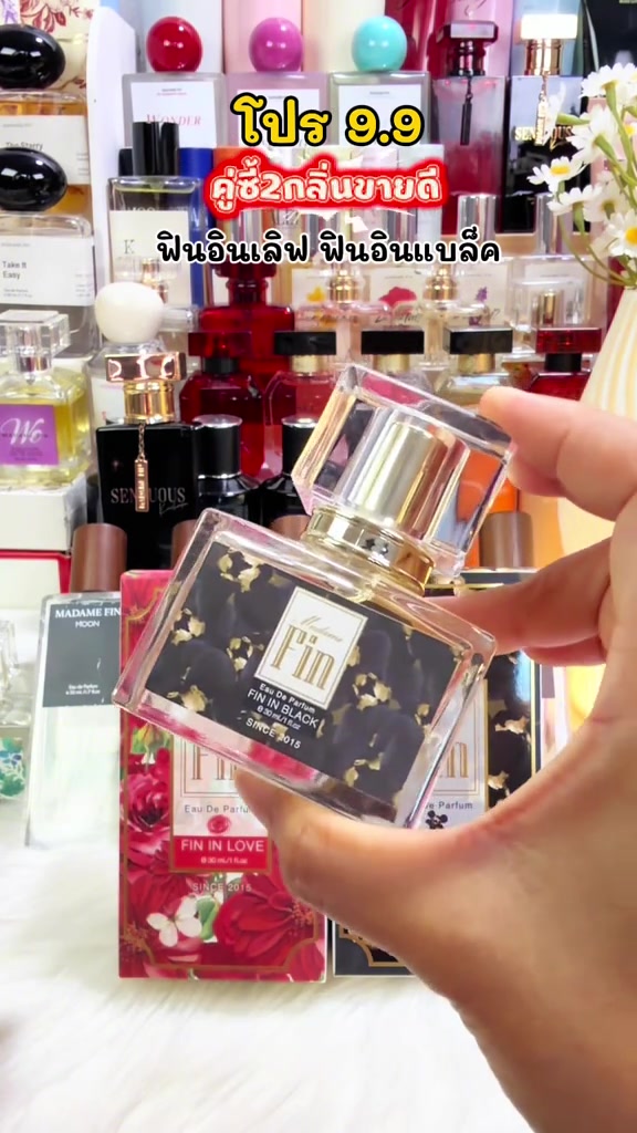 มือถือขวดน้ำหอม Madame Fin Fin in Black ด้านหลังเป็นชั้นวางที่มีขวด WE ฉลากชมพูขวด SENSUOUS สีดำและกล่องน้ำหอมรุ่นอื่นเรียงอยู่