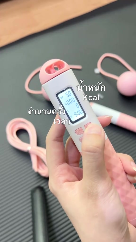 มือถือด้ามเชือกกระโดดอัจฉริยะเห็นหน้าจอแสดงน้ำหนักตัว จำนวนครั้ง เวลา และแคลอรี ฉากหลังเป็นเสื่อสีเทาและลูกตุ้มสีชมพู