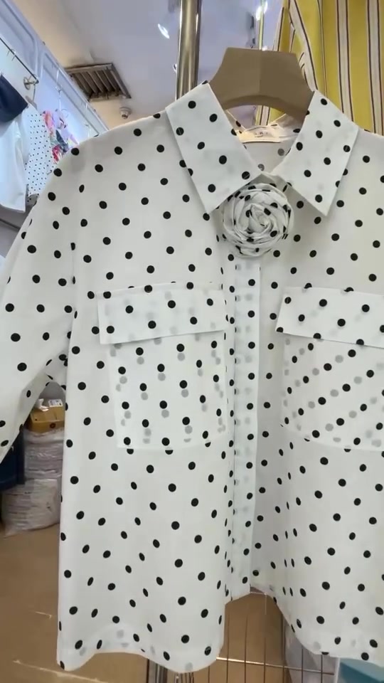 เสื้อเชิ้ตลายจุดดำพื้นขาวแขวนบนไม้แขวนในร้านเสื้อผ้า มีดอกผ้าติดบริเวณปกและกระเป๋าหน้าสองข้าง