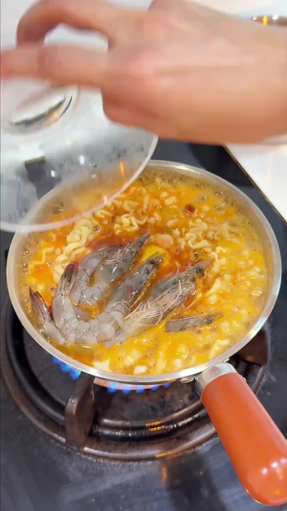 มือเปิดฝาแก้วเหนือหม้อแสตนเลสที่มีกุ้งกับบะหมี่เดือดอยู่บนเตาแก๊ส