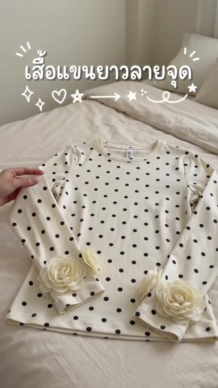 เสื้อแขนยาวลายจุดพื้นครีมวางราบบนผ้าปูเตียงสีอ่อน มีดอกผ้าสีครีมแต่งที่ปลายแขนทั้งสองข้าง