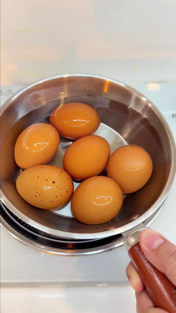 ไข่ไก่หกฟองเรียงอยู่ในหม้อแสตนเลสที่ใส่น้ำ วางบนหน้าเตาสีขาว