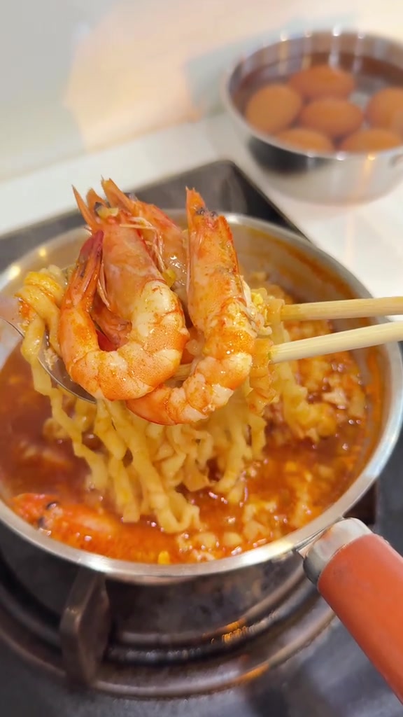 ตะเกียบคีบกุ้งกับเส้นบะหมี่ขึ้นจากหม้อแสตนเลสบนเตา ด้านหลังมีหม้อแสตนเลสอีกใบแช่ไข่ไว้