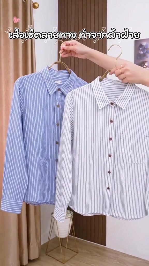 มือถือไม้แขวนเสื้อเชิ้ตแขนยาวลายทางสองตัว ตัวพื้นน้ำเงินทางขาวและตัวพื้นขาวทางน้ำเงิน หน้าฉากม่านสีครีมและผนังไม้