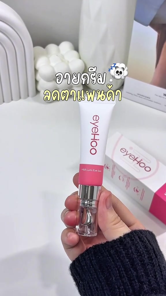 มือถือหลอดอายครีมอายฮูสีขาวแถบชมพู ปลายหลอดเป็นหัวนวดโลหะใส วางคู่กล่องชมพูขาวของอายฮูบนโต๊ะสีขาว