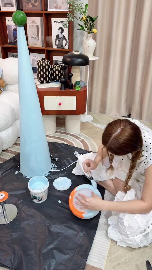 โคมไฟตั้งพื้นทรงกรวยสีฟ้าอ่อนวางข้างถ้วยสีและอุปกรณ์ทาสีบนแผ่นรอง